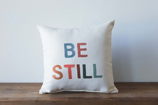 A square poster pillow with the words 'BE STILL' printed in a gradient of colors from blue to red on a beige background.