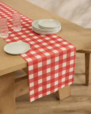 Red and white checkered table runner on a wooden table with plates and glasses. Geometry Ripe Tomato Gingham Table Runner