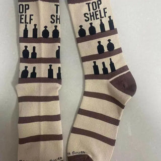 A pair of beige socks with brown stripes and graphics of bourbon bottles, text 'TOP SHELF', and the brand 'Barrel Down South'.