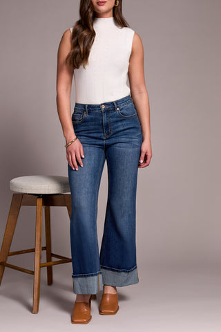 Woman wearing a white sleeveless top and blue jeans standing next to a wooden stool with a gray cushion.