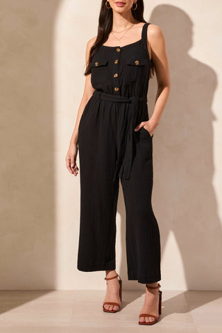 A sleeveless black jumpsuit with button-front bodice, tank top straps, and patch pockets, worn by a model standing against a light background.