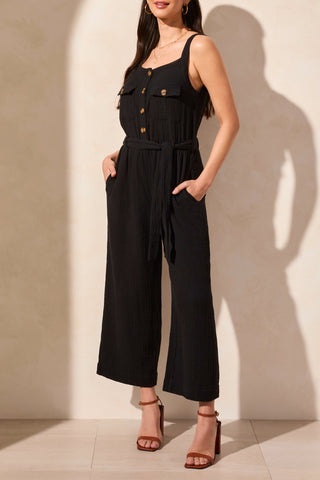 A sleeveless black jumpsuit with button-front bodice, tank top straps, and patch pockets, worn by a model standing against a light background.