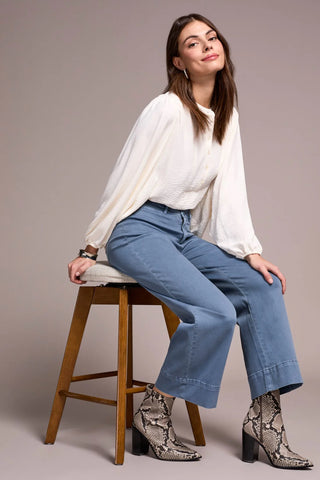 Woman sitting on a stool wearing a white blouse, blue jeans, and patterned shoes against a plain background
