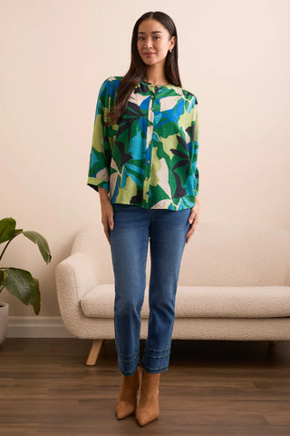 Woman wearing a colorful blouse and blue jeans standing in a room with a beige wall and a white chair.