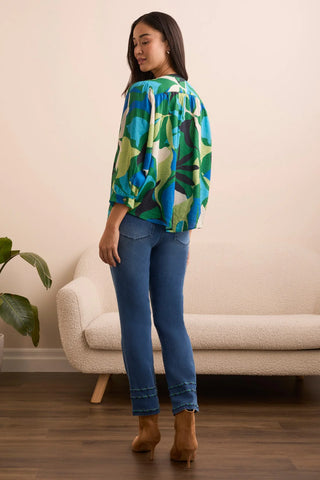 Woman wearing a colorful patterned shirt and blue jeans standing in a room with a beige sofa and plant.