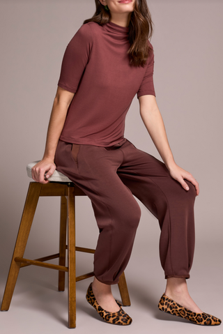 Woman sitting on a wooden stool wearing a brown outfit with leopard print shoes against a plain background