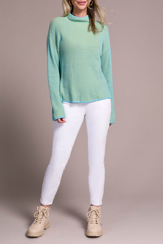 Person wearing a light green and blue sweater and white pants against a plain background