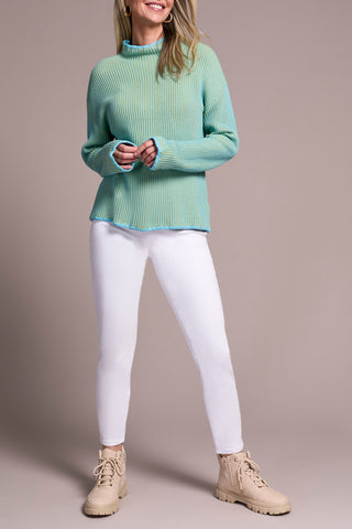 Woman wearing a light green and blue sweater, white pants, and beige boots against a plain background