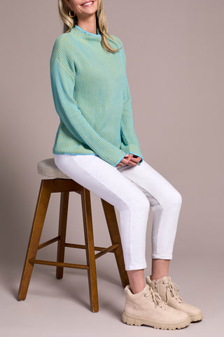 Woman sitting on a wooden stool wearing a green and blue sweater, white pants, and beige boots against a plain background