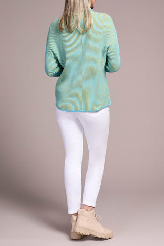 Person wearing a blue and green sweater, white pants, and beige boots on a plain background