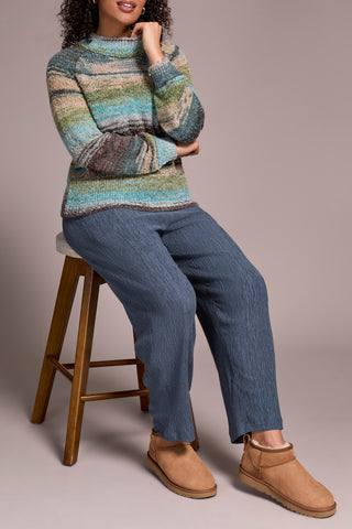 Woman wearing a striped sweater and blue jeans sitting on a wooden stool against a plain background