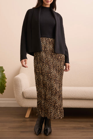 Woman wearing a black jacket, black top, and leopard print skirt in a room with a beige sofa and plant.