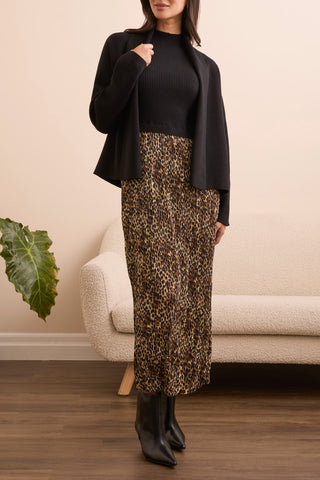 Person wearing a black top, black jacket, and leopard print skirt standing in a room with a beige wall and a plant.