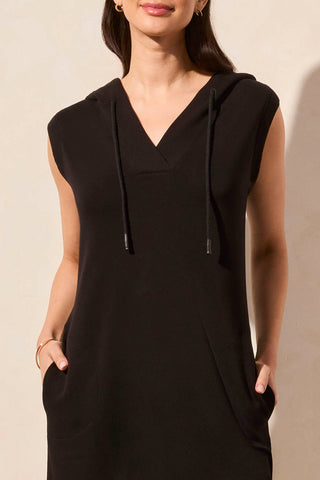 A woman standing wearing a black sleeveless hooded dress with pockets and a V-neckline.