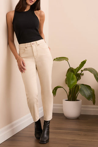 Woman wearing beige pants and black boots standing next to a potted plant indoors.