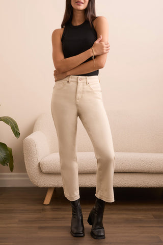Woman wearing beige jeans and black top standing in a room with a beige armchair and plant.