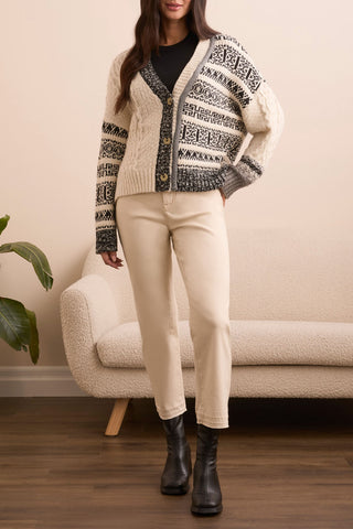 Person wearing a patterned cardigan, black shirt, white pants, and black boots in a room with a beige sofa and plant.