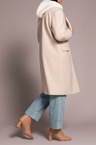 Person wearing a beige coat, light blue jeans, and brown shoes on a plain background