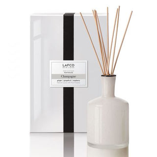 A LAFCO champagne-scented diffuser with a group of wooden reeds in a white vase, in front of a black and white box with the product label.