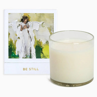 A candle with a white wick, placed in a clear glass holder, with a card behind it featuring an artwork of an angel in a field of green and yellow. Anne Neilson Be Still Candle