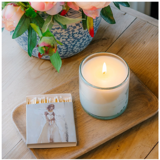 A hand-poured white candle in an artisan glass, with a card beside it featuring a painting of a angel. The candle is labeled 'GRACE', Anne Neilson Grace Candle