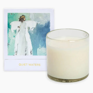 A hand-poured white candle next to its packaging which features artwork by Anne Neilson. The candle is in a clear glass container and there are words 'QUIET WATERS' visible on the packaging.