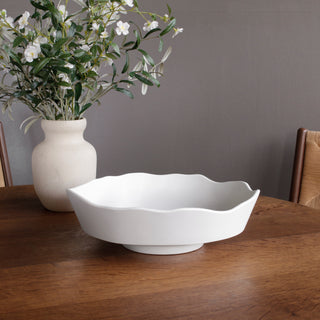 A white, melamine Dakota large bowl with a chiseled rim and faceted edge on a raised base, displayed on a wooden table with a vase of flowers in the background. Beatriz Ball Vida Nube Dakota Large Bowl, White
