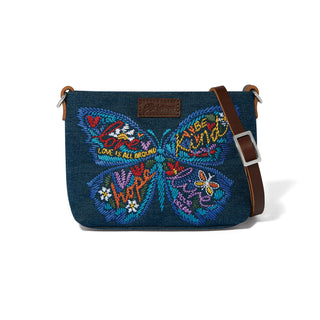 A dark blue denim crossbody bag featuring a large colorful embroidered butterfly on the front with the words 'Love is all around' and 'Keep it kind'. The bag has brown leather trim and a brown adjustable shoulder strap with a silver buckle, set against a plain white background. Brighton Dream Wings Denim Embroidered Pouch