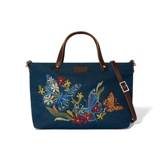 A navy blue denim tote bag with brown leather handles and a matching shoulder strap, featuring colorful embroidered butterflies and flowers on the front. Brighton Love Flutter Denim Embroidered Tote