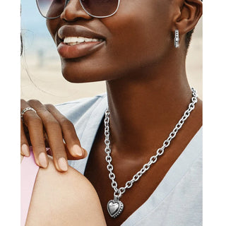Woman wearing a silver chain necklace with a heart pendant, earrings, and sunglasses. Brighton Pretty Tough Bold Heart Necklace