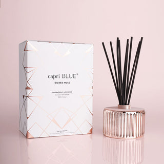 A rose gold Reed diffuser by Capri Blue with black sticks, placed next to its white and gold box packaging.