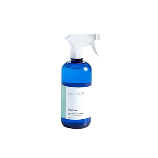 A blue bottle with a white spray nozzle labeled 'Capri Blue Volcano Multi-Surface Cleaner' on a light blue background.