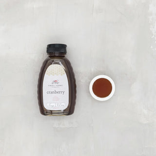 A bottle of Cranberry Honey with a black cap and a small white dish of the honey next to it, placed on a neutral background, finch and fennel