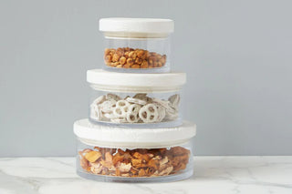 A set of clear glass canisters with white lids, stacked on top of each other, filled with what appears to be snacks like pretzels and nuts. ETU