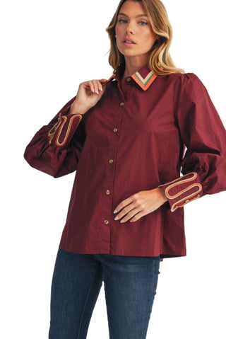 Woman wearing a burgundy blouse with decorative cuffs and collar against a beige background. Embroidery Accent Button Down Shirt in Burgundy