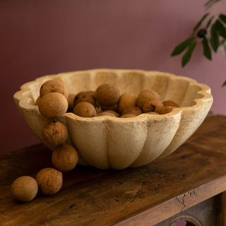 A beige faux travertine ruffled bowl filled with round decorative items, placed on a wooden surface against a pink wall.