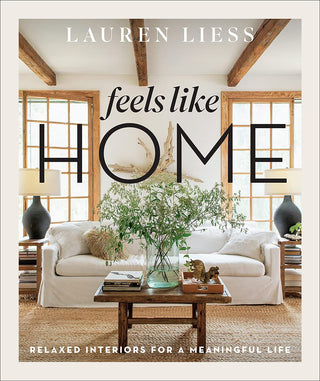 The cover of the book 'Feels Like Home' by Lauren Liess featuring a photograph of a cozy living room with a white sofa, wooden beams on the ceiling, and a large window. Feels Like Home.