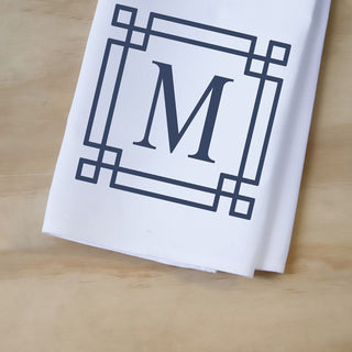 White towel with a black geometric border and letter 'M' on a wooden surface