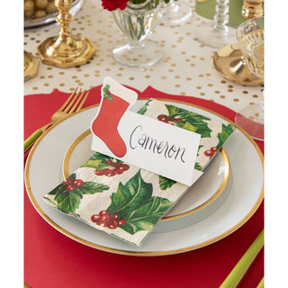 Christmas-themed place setting with holly design napkin and name card on a red tablecloth.