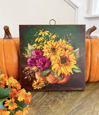 Jeweled Flower Pumpkin Large Art
painting on metal depicting a vibrant floral arrangement with yellow sunflowers, purple roses, and black-eyed flowers in a pumpkin-shaped vase against a black and green background. The painting is shown hanging from a metal ring at the top.