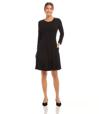 A woman standing in a studio setting, wearing a knee-length black dress with 3/4 sleeves, scoop neck, and fit and flare silhouette. 