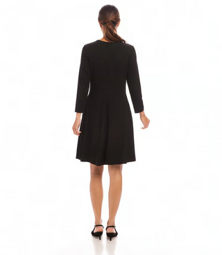 A woman standing in a studio setting, wearing a knee-length black dress with 3/4 sleeves, scoop neck, and fit and flare silhouette. 