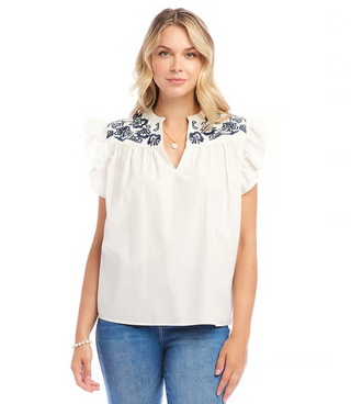 A woman wearing an off white top with navy blue embroidered ruffle sleeves and a split neck.