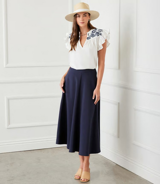 A woman wearing an off white top with navy blue embroidered ruffle sleeves and a split neck.