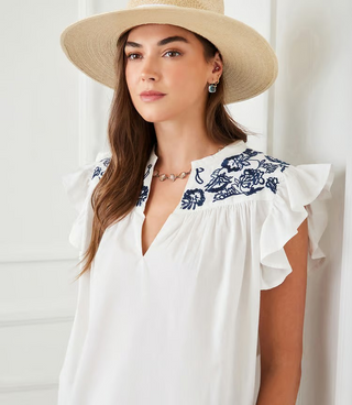 A woman wearing an off white top with navy blue embroidered ruffle sleeves and a split neck.
