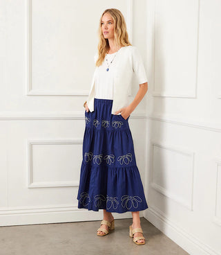 A woman wearing an off-white short sleeve ribbed cardigan sweater with a V-neck, paired with a blue skirt with decorative white detail.