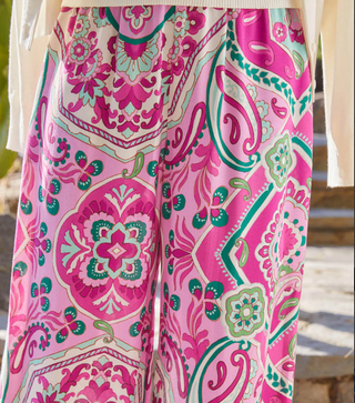 A pair of high-waisted, wide-leg pants with a paisley print in pink and green hues on a crepe fabric. They feature a pull-on elastic waistband