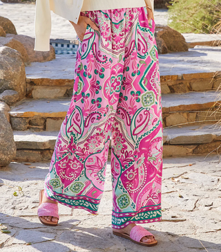 A pair of high-waisted, wide-leg pants with a paisley print in pink and green hues on a crepe fabric. They feature a pull-on elastic waistband