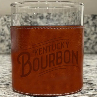 Glass with a brown leather sleeve displaying 'Kentucky Bourbon' on a marble surface. Kentucky Bourbon Word Angled Faux Leather Rocks Glass