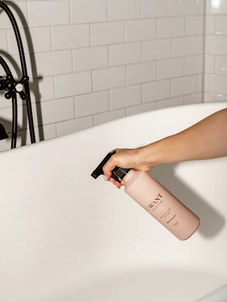 Person holding a pink bottle with a black nozzle over a bathtub in a bathroom setting. L'AVANT Blushed Bergamot Multipurpose Cleaner